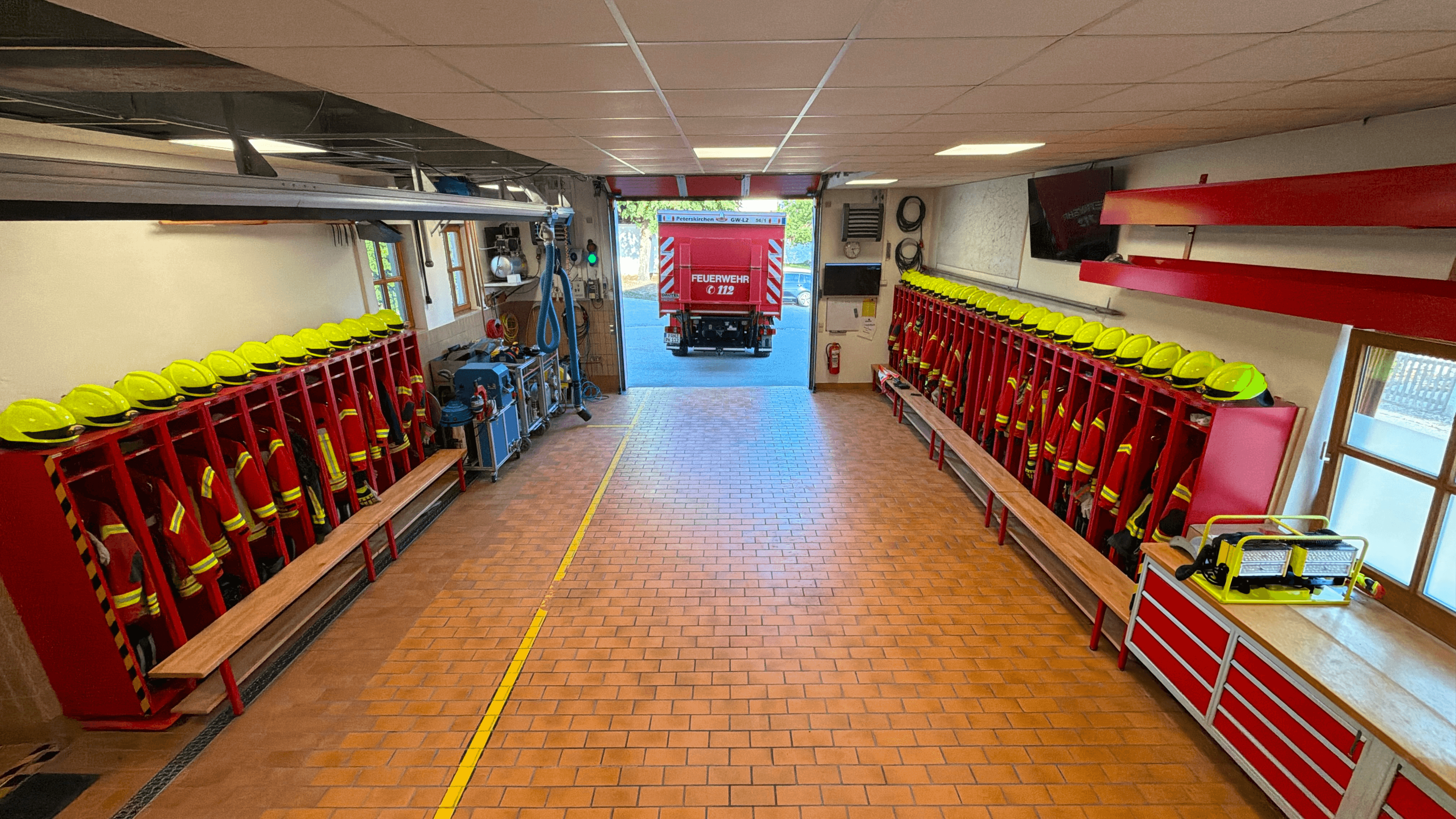 Fahrzeughalle des Feuerwehrhauses mit Spinden auf beiden Seiten, im Hintergrund das ausfahrende Feuerwehrfahrzeug vor dem geöffneten Sectionaltor